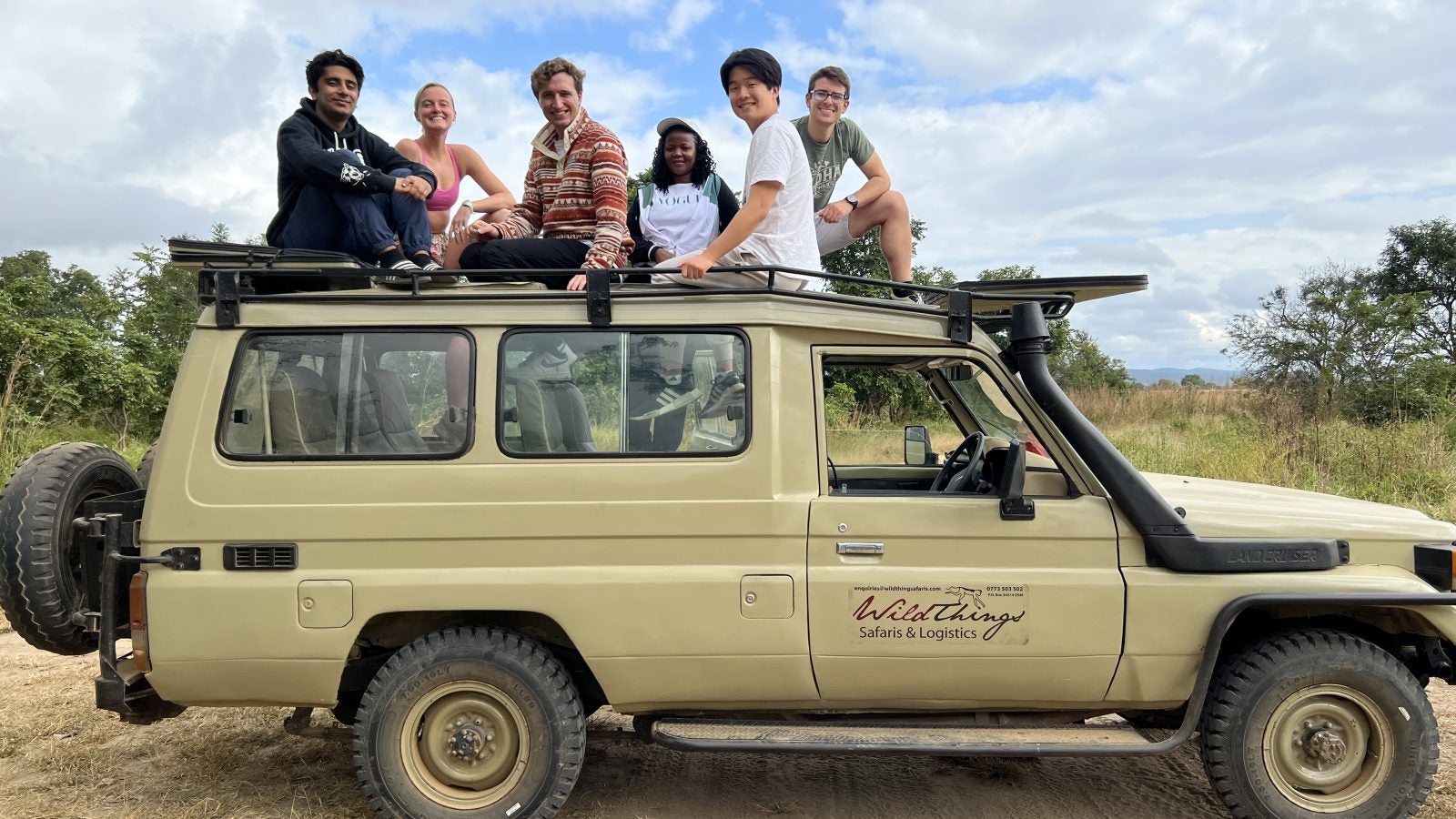 Group of tourists sitting on and standing around a safari vehicle in a savannah landscape.