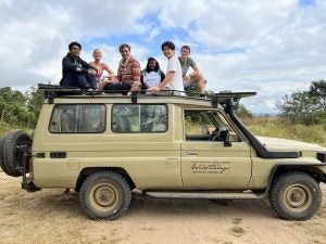 Group of tourists sitting on and standing around a safari vehicle in a savannah landscape.