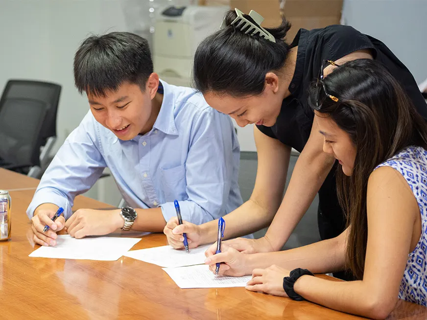 Two students and a faculty member, leaning on a table, writing on papers