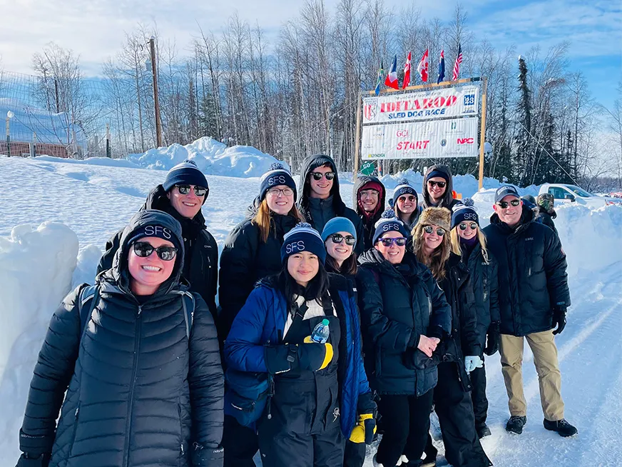 Group of students in the snow in Alaska, behind them a sign for a sled dog race