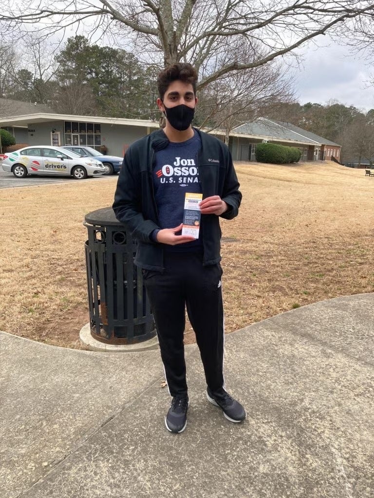 Arash Abbas stands in front of a parking lot. He is wearing a face mask, a Jon Ossoff for Senate T-shirt and is holding an Ossoff campaign leaflet.