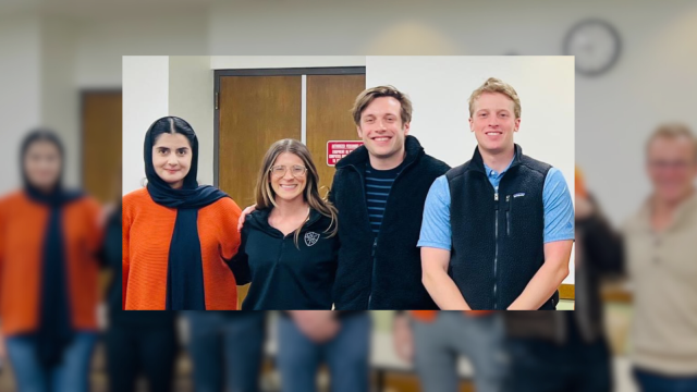 Group photo of seven individuals smiling in an indoor setting with a clock on the wall.