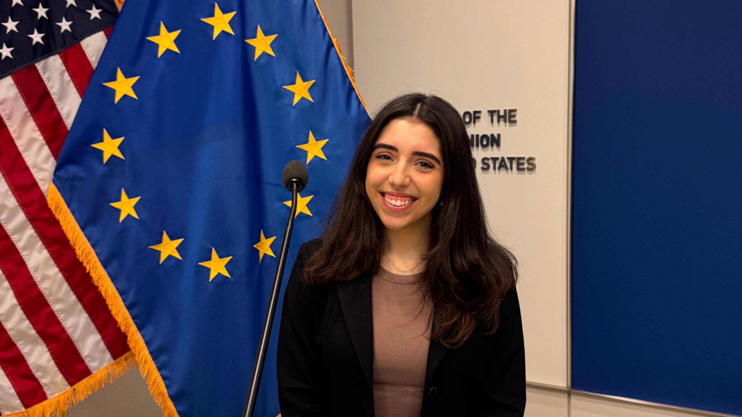 A woman with long dark hair wearing a black blazer stands in front of the European Union and American flags at a podium.