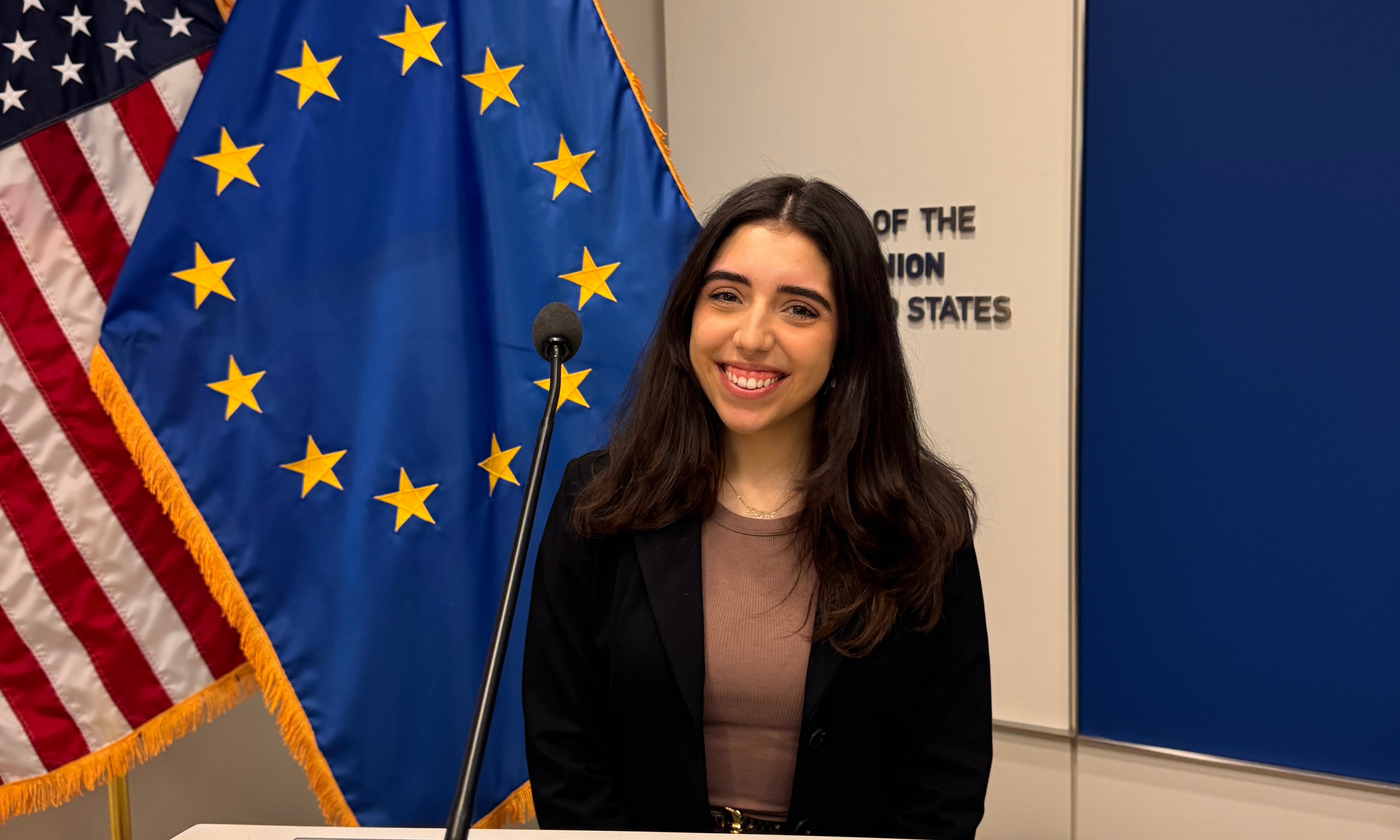 A woman with long dark hair wearing a black blazer stands in front of the European Union and American flags at a podium.