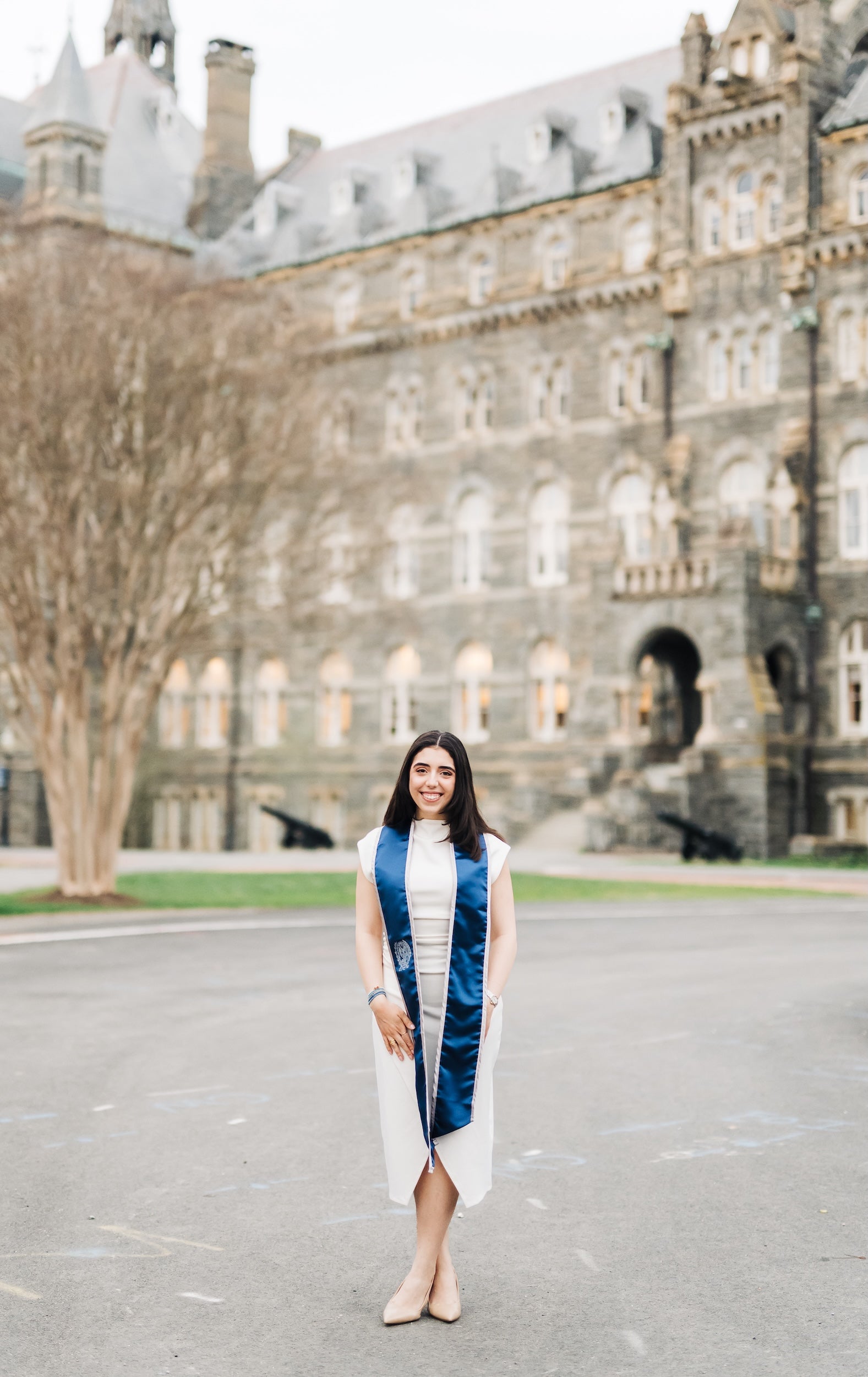 A woman with dark hair wearing a white dress and blue graduation stole stands in front of Healy Hall.