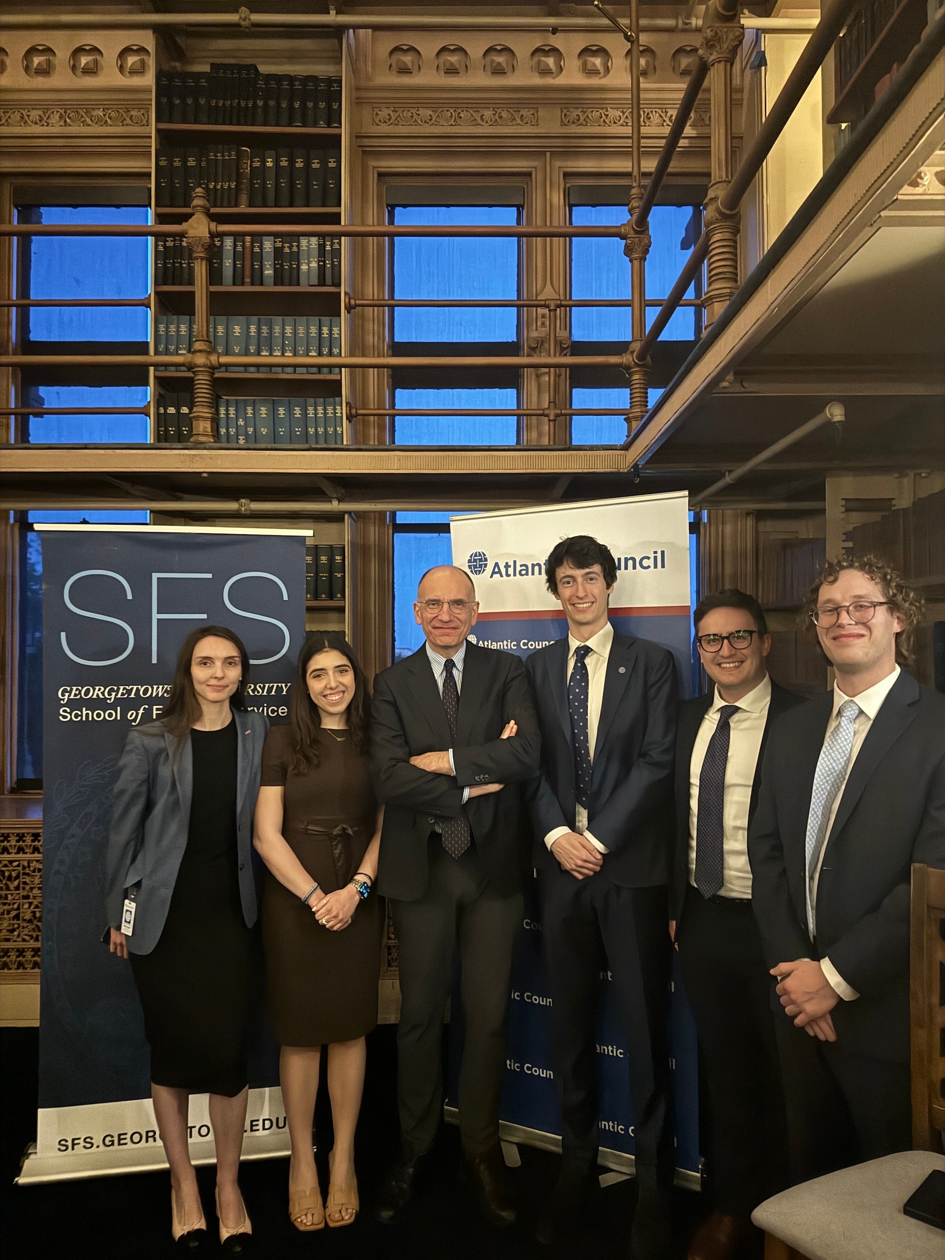 Five individuals posing together at an event hosted by Georgetown University's School of Foreign Service and the Atlantic Council, standing in front of promotional banners inside a room with traditional wooden bookshelves.