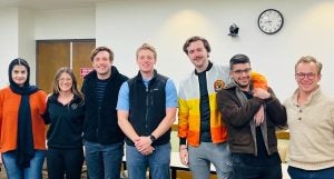 Group photo of seven individuals smiling in an indoor setting with a clock on the wall.
