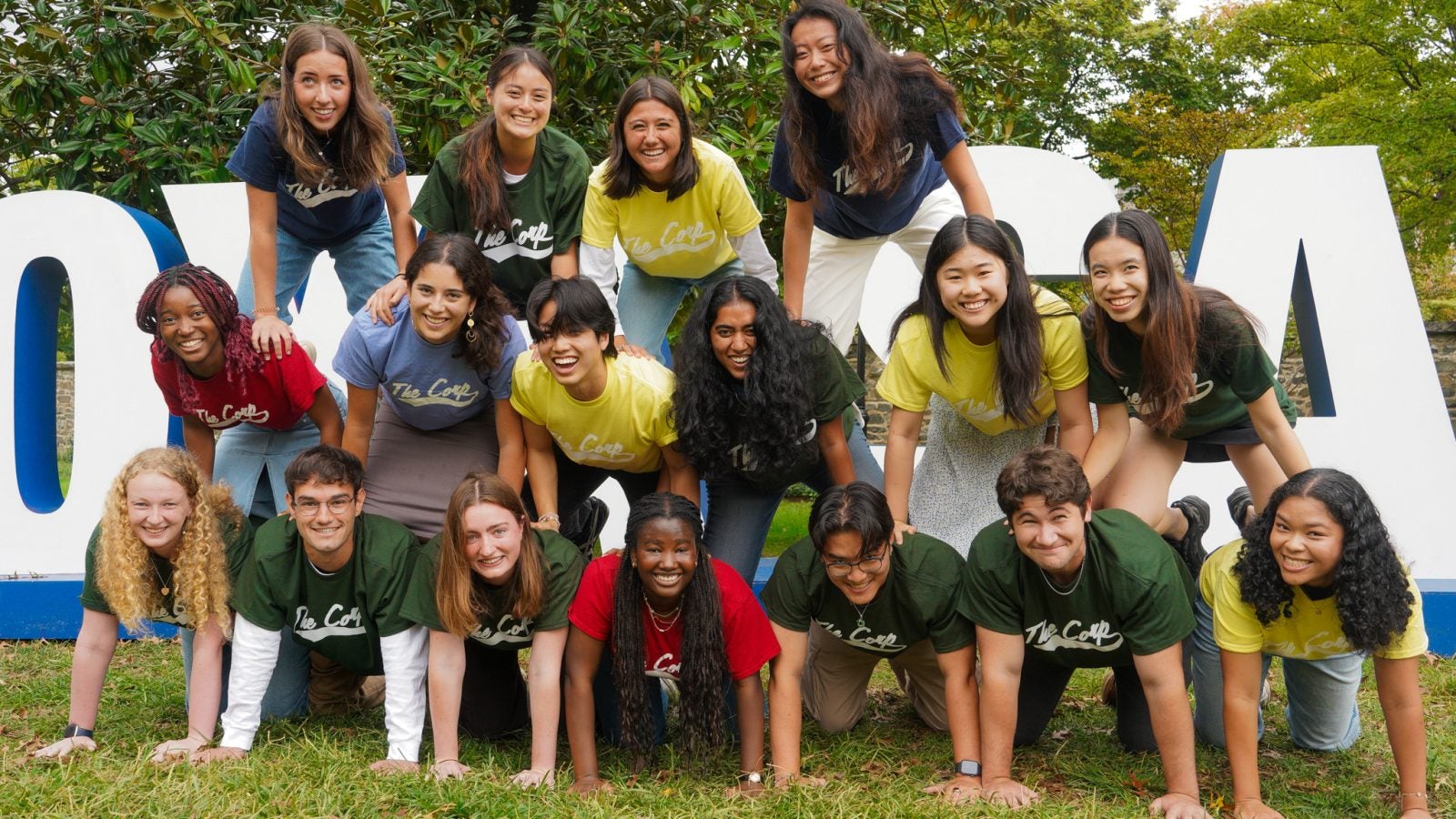 Group of students wearing matching t-shirts posing playfully around large &quot;HOYA SAXA&quot; sign outdoors.
