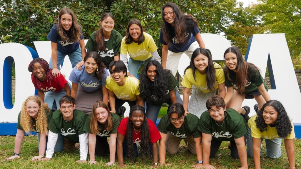 Group of students wearing matching t-shirts posing playfully around large "HOYA SAXA" sign outdoors.