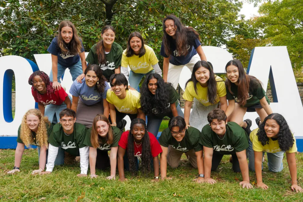 Group of students wearing matching t-shirts posing playfully around large "HOYA SAXA" sign outdoors.