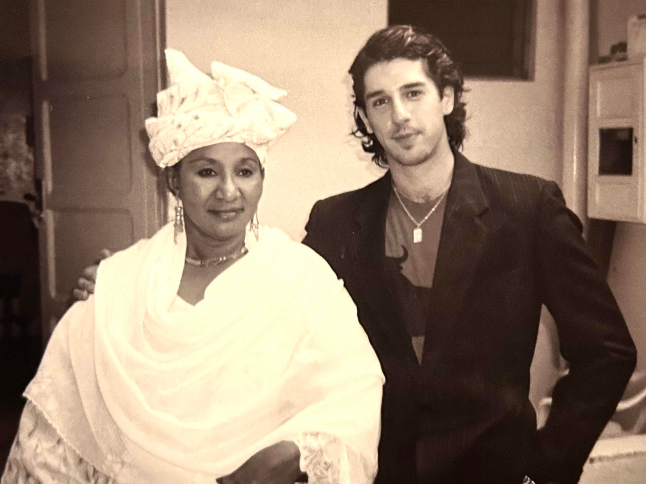 A woman dressed in a traditional white outfit with headwrap standing next to a man with long dark hair wearing in a dark suit.