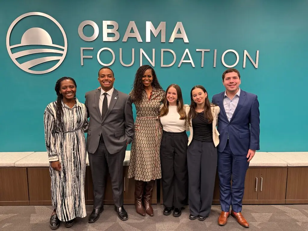 Group of six people posing for a photo in a professional setting, including Mrs. Michelle Obama. the Obama Foundation logo is displayed on a blue wall in the background.