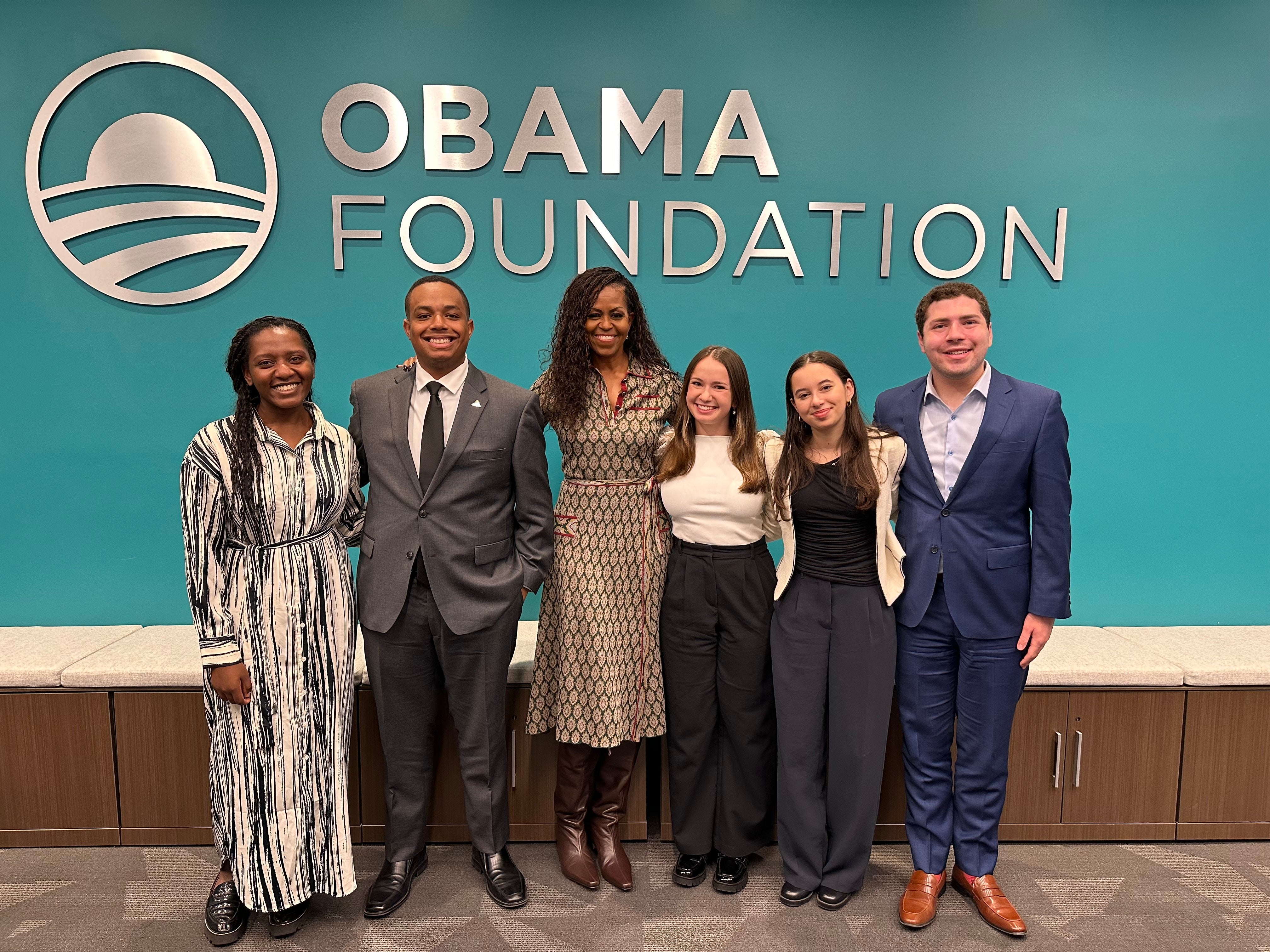 Group of six people posing for a photo in a professional setting, including Mrs. Michelle Obama. the Obama Foundation logo is displayed on a blue wall in the background.
