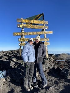 Two people standing at the summit of Mount Kilimanjaro next to a congratulatory signboard indicating Uhuru Peak, Tanzania, at an altitude of 5895 meters.