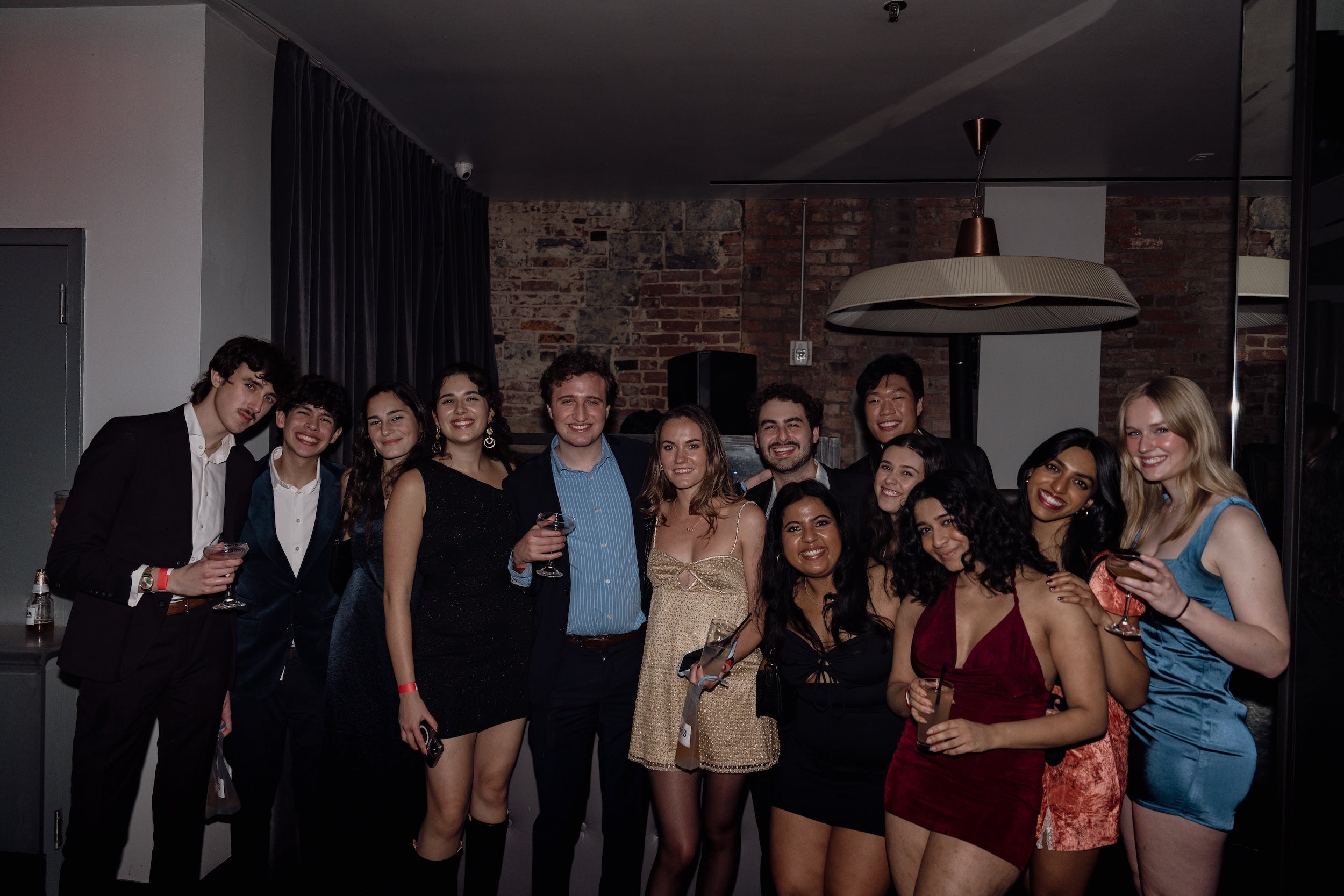Group of young adults smiling and posing together at a party, some holding drinks. They are dressed in semi-formal attire.