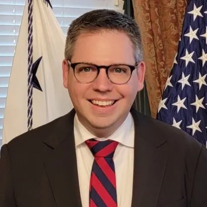 Eric Jacobstein wearing a blazer and a red and navy striped tie and glasses. Two flags are in the background.