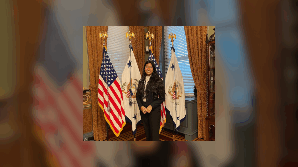 A person standing between two U.S. flags and a flag with a blue and white emblem in a formal office setting.
