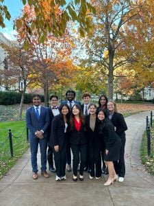 A group of nine people dressed in business attire smiling and standing outdoors with autumn foliage in the background.
