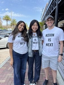 Three people wearing matching white t-shirts with the text "KEVIN IS NEXT" printed on them, standing together smiling on a sunny, tree-lined street.
