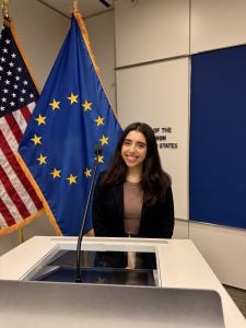 Sophia Athan stands in front of the European Union and American flags at a podium.