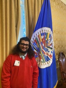 Person standing next to the Organization of American States flag in an elegant room.