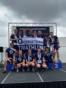 Group of athletes from Georgetown Triathlon team celebrating on a podium with medals, holding a banner with the team name.