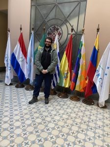 A person smiling and standing in front of a collection of international flags in a tiled hallway.