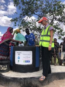 Group of individuals drawing water from a well marked by a sign indicating sponsorship by WF-AID, set in a rural landscape under a clear sky.