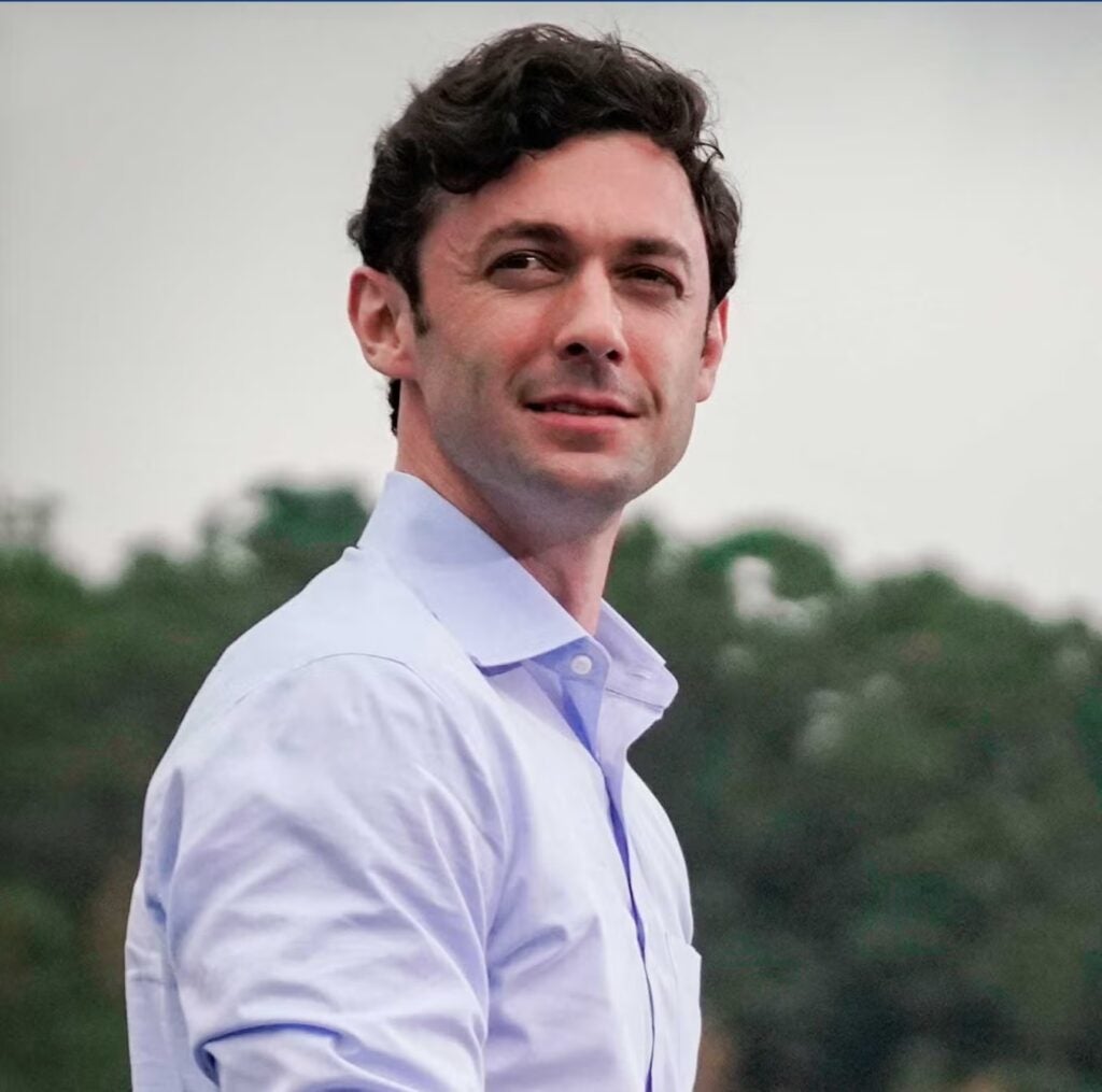 Senator Jon Ossoff standing, wearing a light blue shirt. Behind him green trees