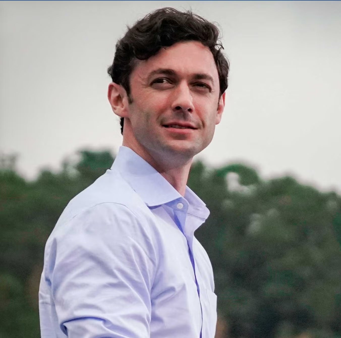 Senator Jon Ossoff standing, wearing a light blue shirt. Behind him green trees