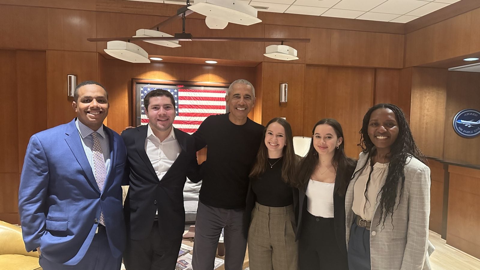 Former President Barack Obama stands smiling in the center of a group of five diverse professionals in a boardroom, with an American flag in the background.