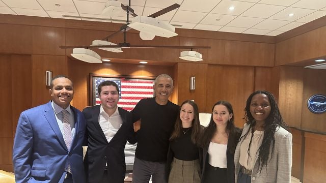 Former President Barack Obama stands smiling in the center of a group of five diverse professionals in a boardroom, with an American flag in the background.