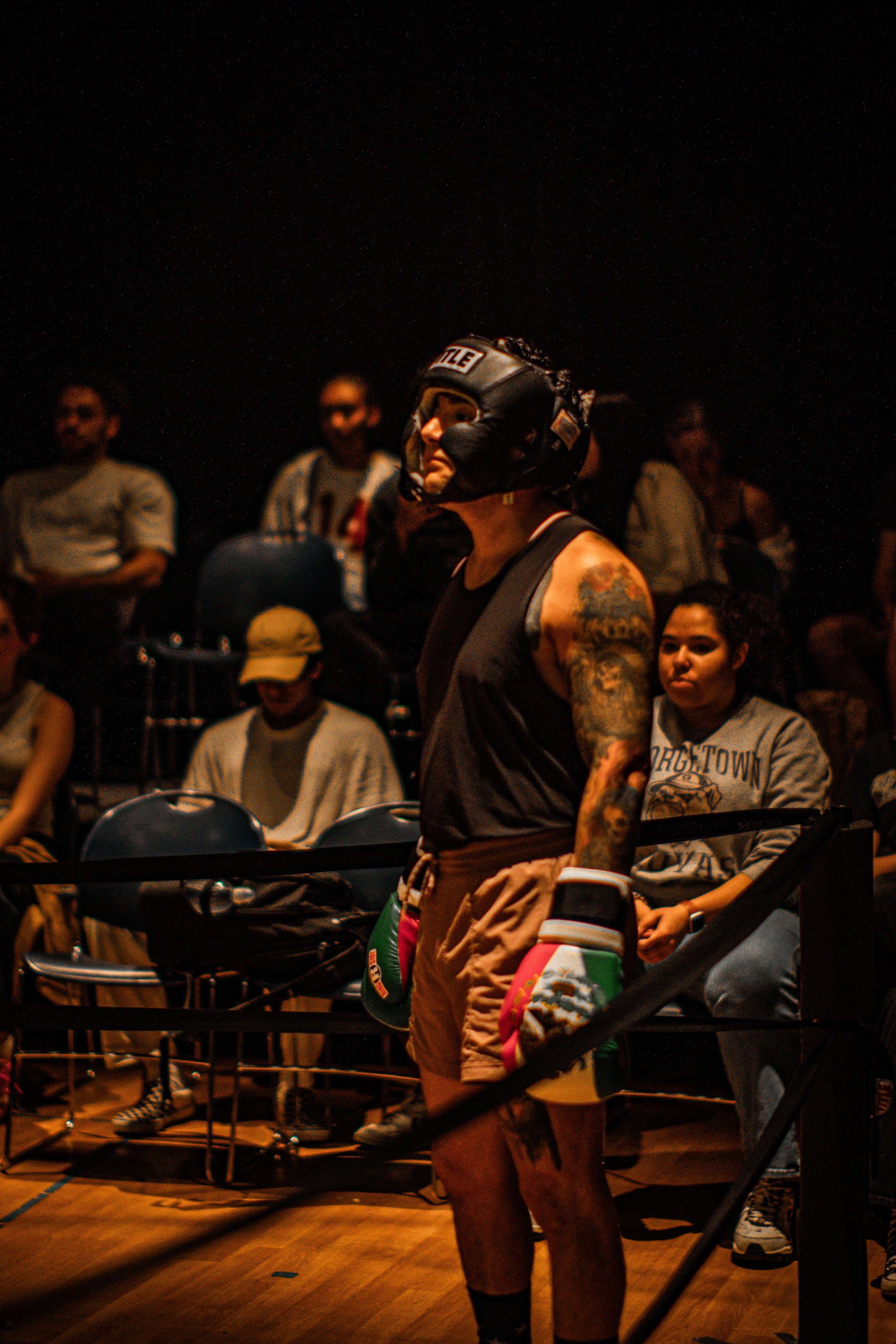 Person in boxing gear with headgear and gloves stands confidently in a dimly lit arena, surrounded by spectators.