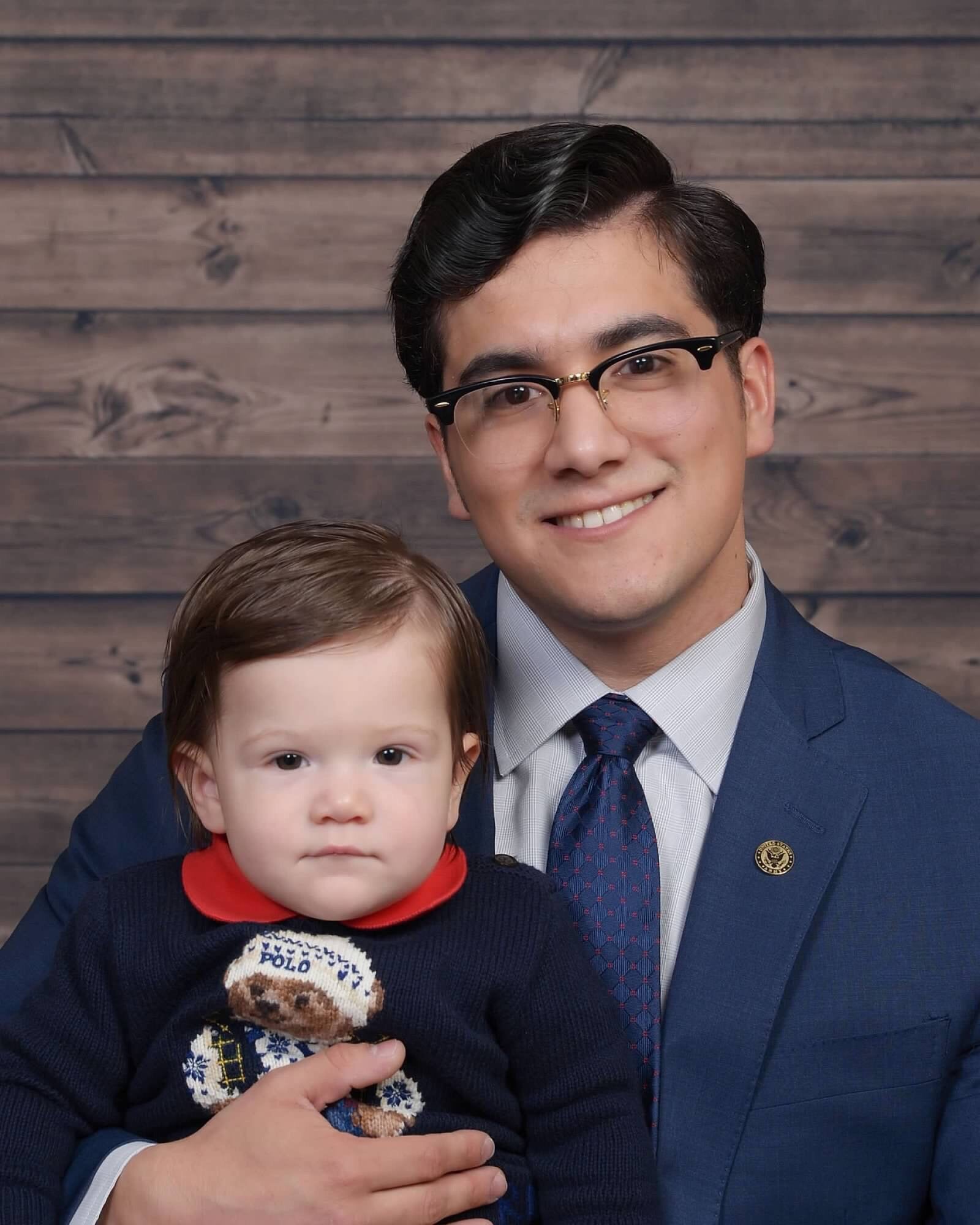 An individual in a suit and glasses holding a small child dressed in a sweater with a teddy bear design, both smiling against a wooden plank backdrop.