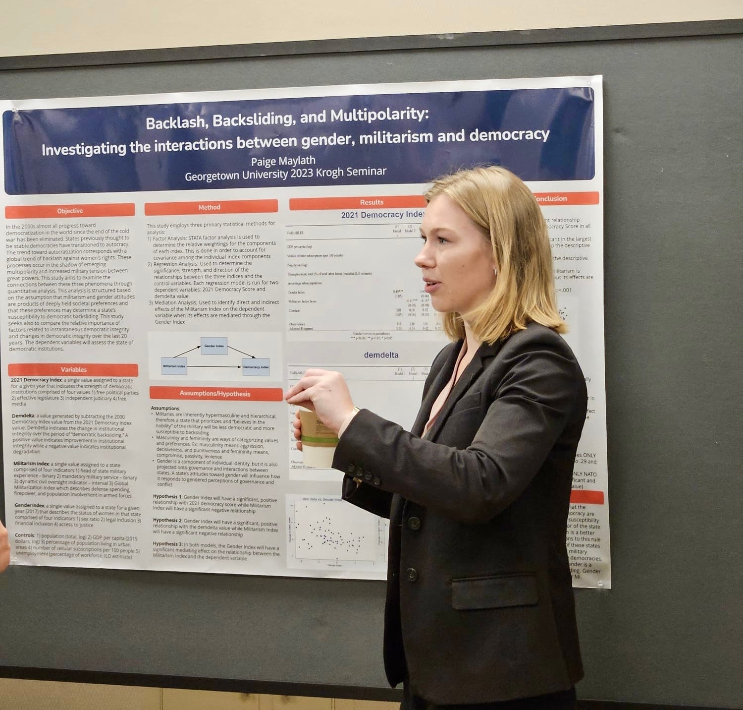 Paige Maylath, a blond woman in a dark suit, stands in front of a poster titled "Backlash, Backsliding and Multipolarity"