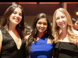 Three people smiling at a formal event, standing closely together. They are dressed in elegant attire.