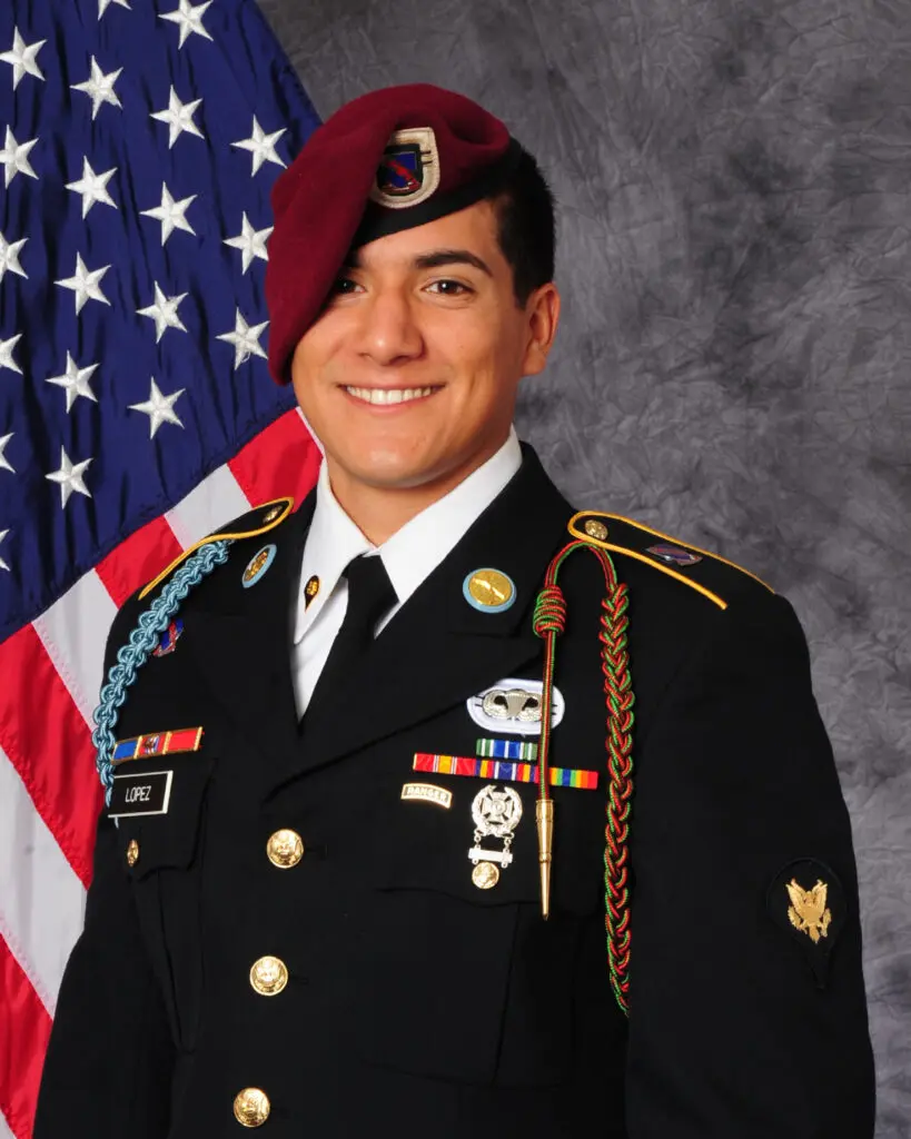 A U.S. Army soldier in dress uniform, with various medals and a maroon beret, posing in front of an American flag.