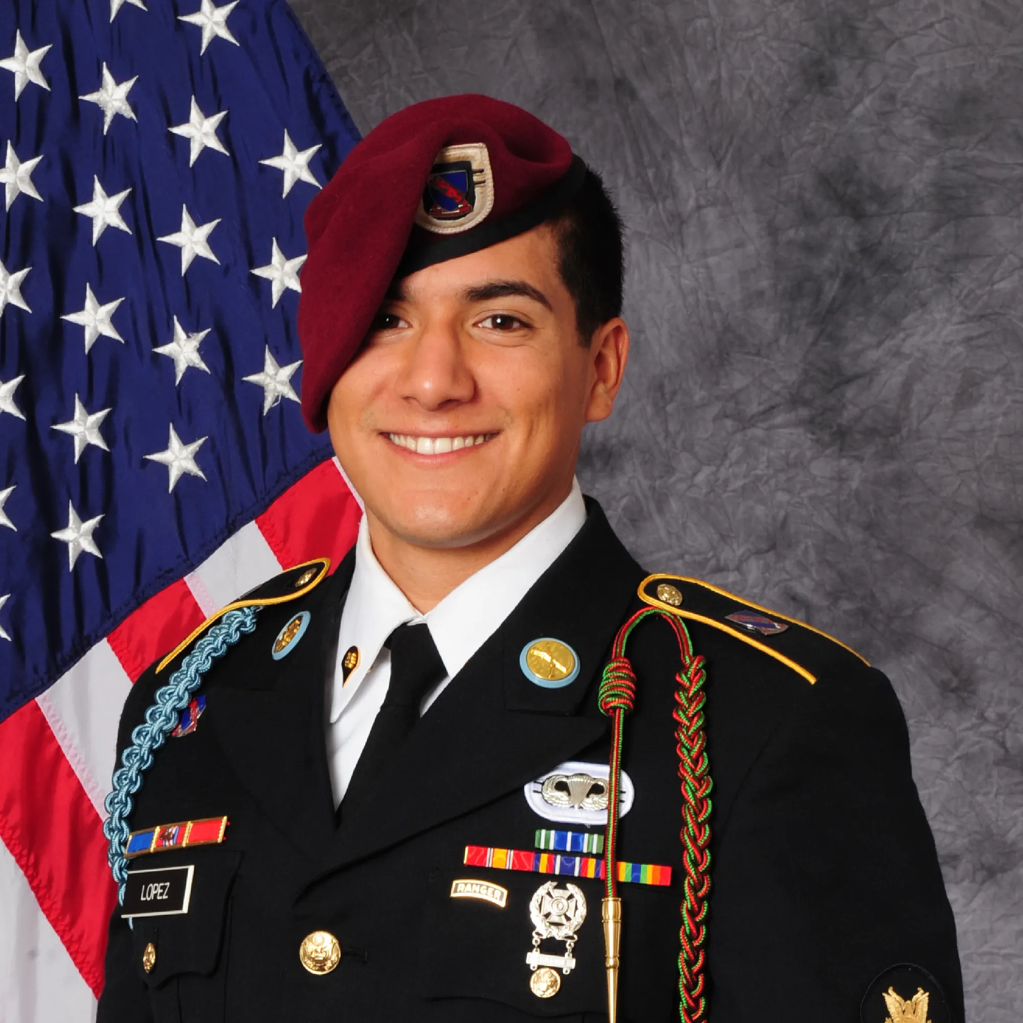 A U.S. Army soldier in dress uniform, with various medals and a maroon beret, posing in front of an American flag.
