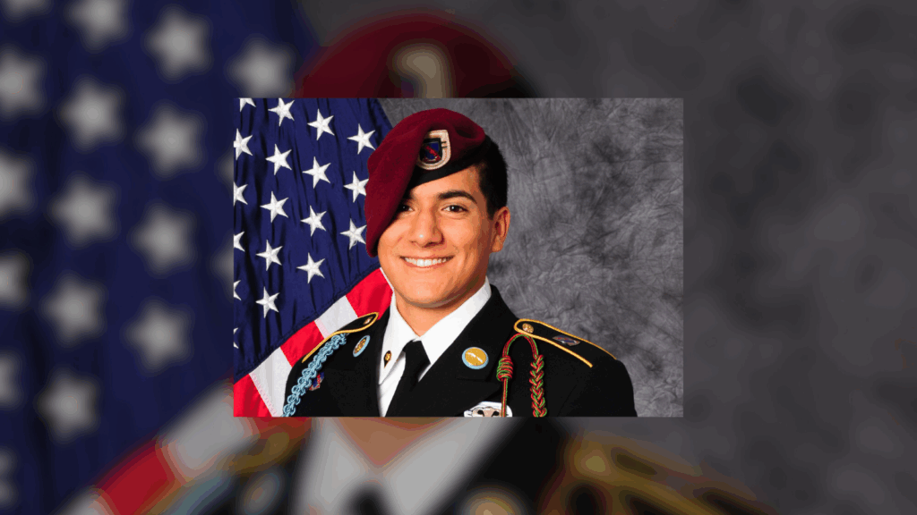 A U.S. Army soldier in dress uniform, with various medals and a maroon beret, posing in front of an American flag.