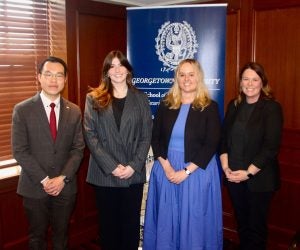 Four individuals smiling and standing in a row in front of a Georgetown University banner in a wood-paneled room. They are dressed in professional attire.