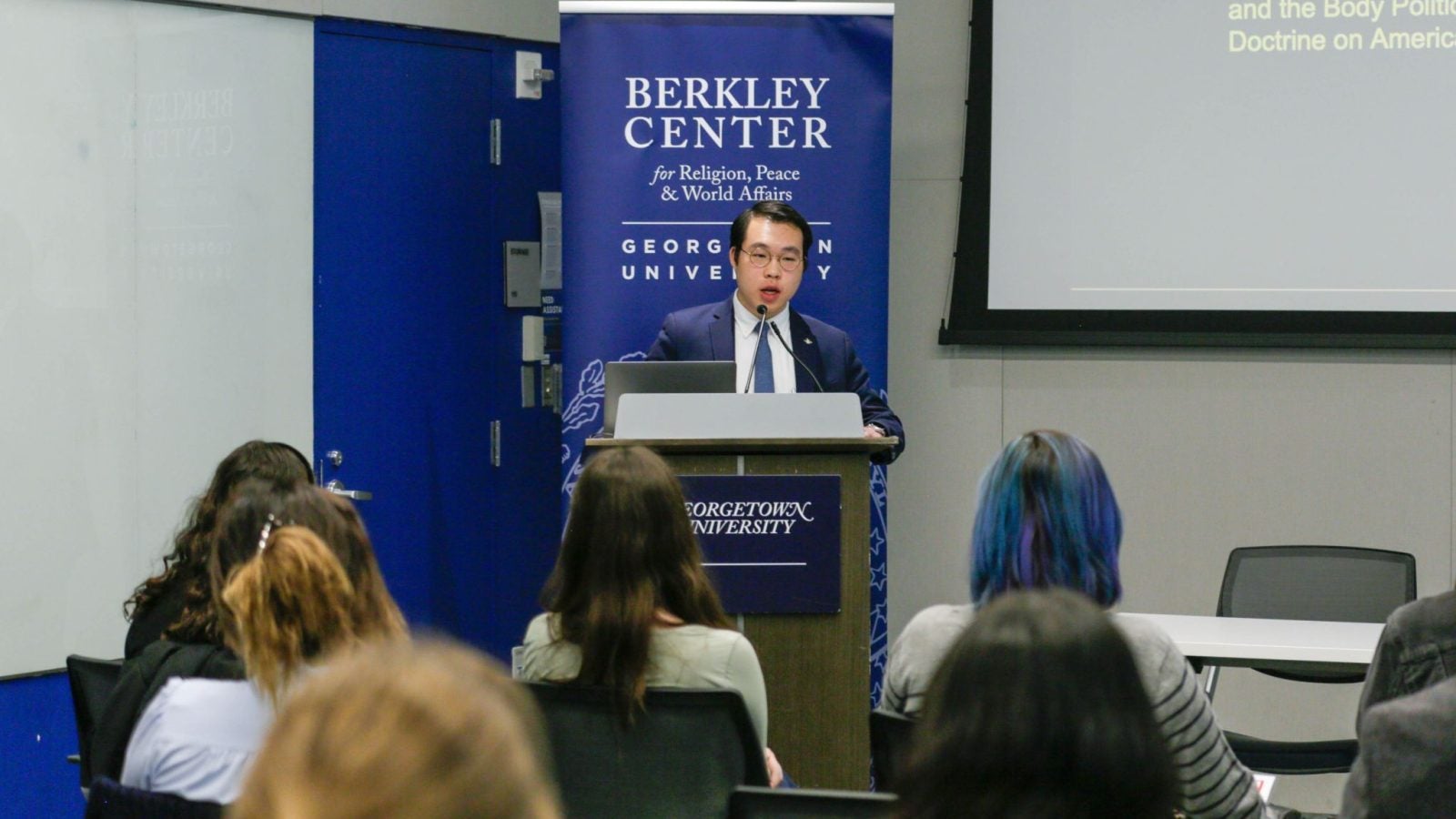 An individual speaks at a podium, in a room with attendees and banners labeled &quot;Berkley Center for Religion, Peace &amp; World Affairs.
