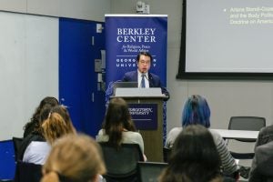 An individual speaks at a podium, in a room with attendees and banners labeled "Berkley Center for Religion, Peace & World Affairs.