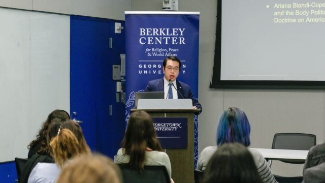 An individual speaks at a podium, in a room with attendees and banners labeled &quot;Berkley Center for Religion, Peace &amp; World Affairs.