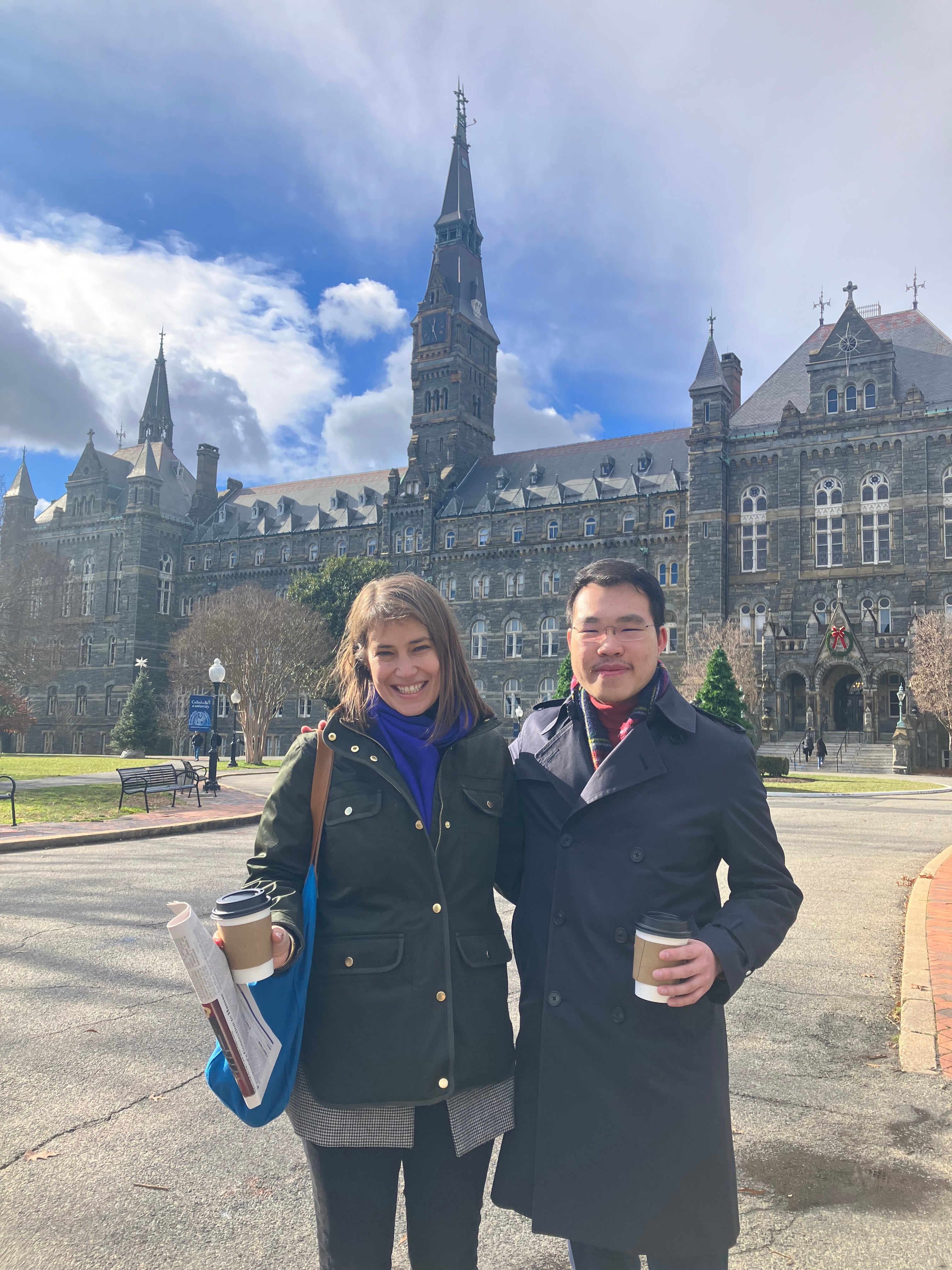 Two people smiling in front of Healy Hall at Georgetown University, one holding a coffee cup and the other carrying books and a backpack.