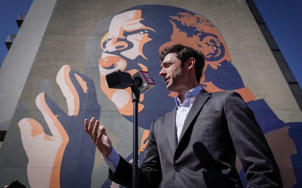 Ossoff speaking in front of a microphone. Behind him a wall with portrait of a man painted in blue, pink and orange.