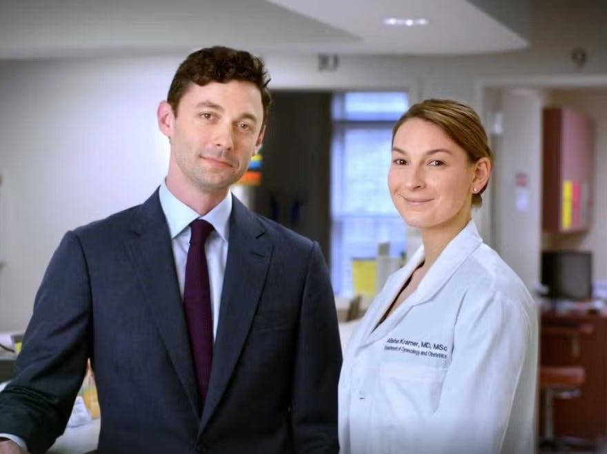 Senator Jon Ossoff standing next to his wife Alisha Kramer. He is wearing a suit, she is wearing a labcoat.