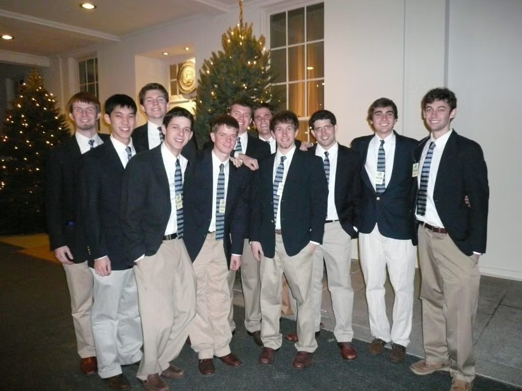 A group of 12 young men wearing navy jackets, and khaki pants, posing for a photograph. 