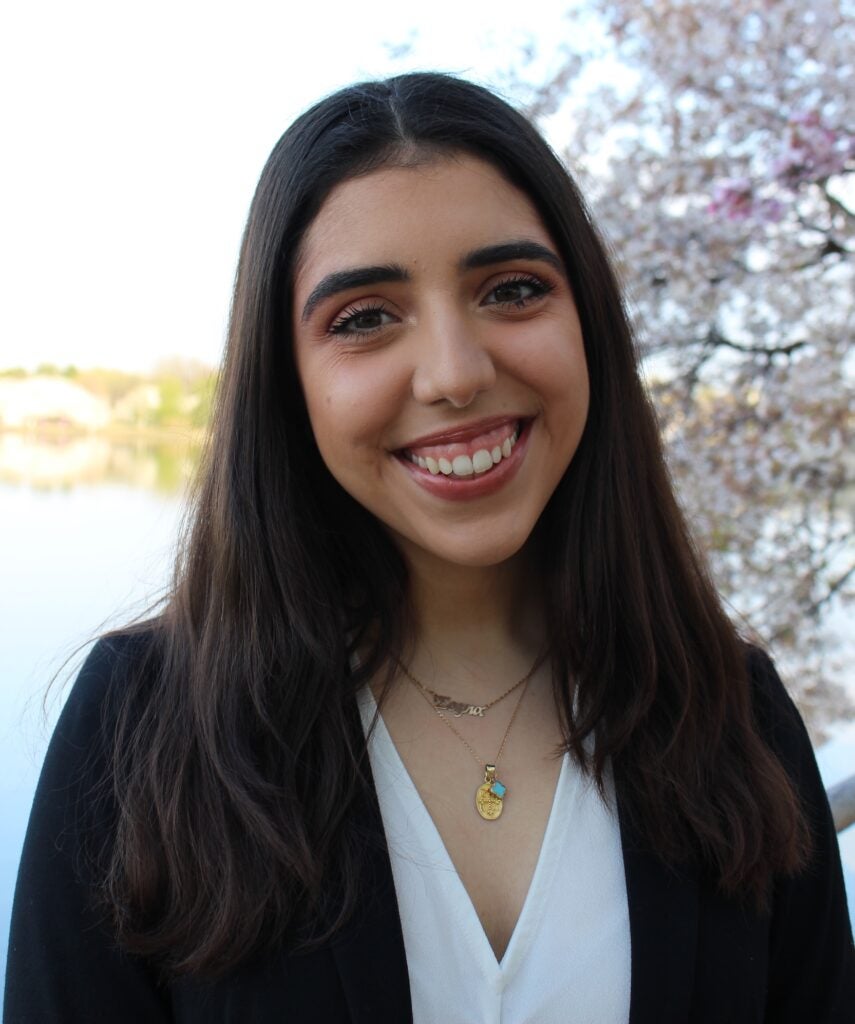 Individual smiling, standing outdoors with a lake and flowering trees in the background.