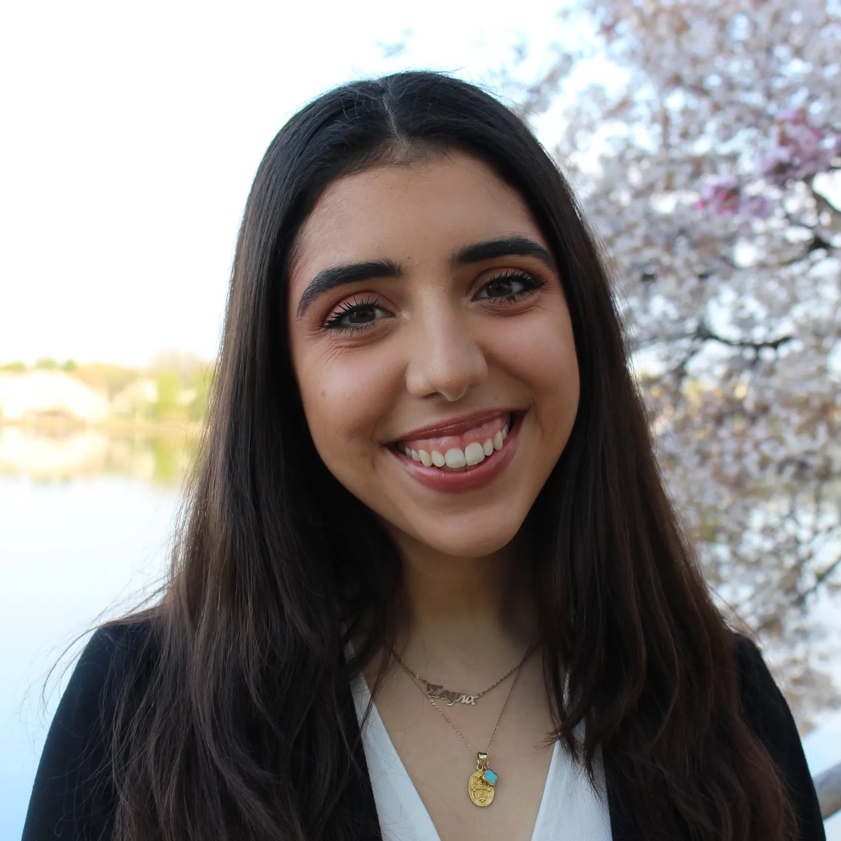 Individual smiling, standing outdoors with a lake and flowering trees in the background.