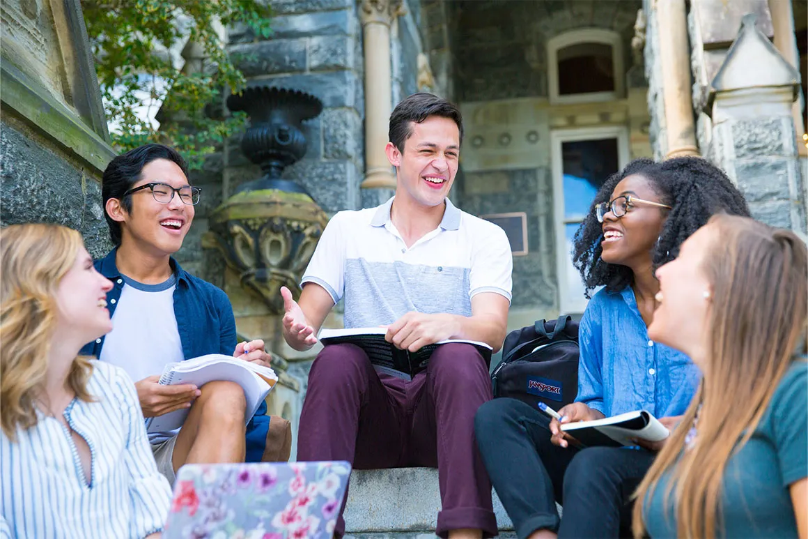 Five students sitting on Healy steps, laughing with books on their laps.